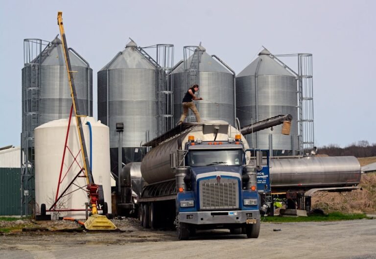 A worker loads a fuel truck in front of large industrial silos outdoors.